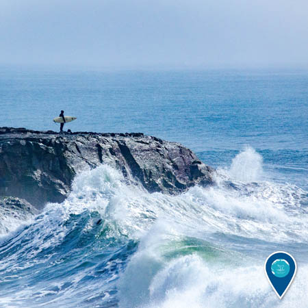 surfer standing on a rock cliff looking out at the ocean while wave crash round him
