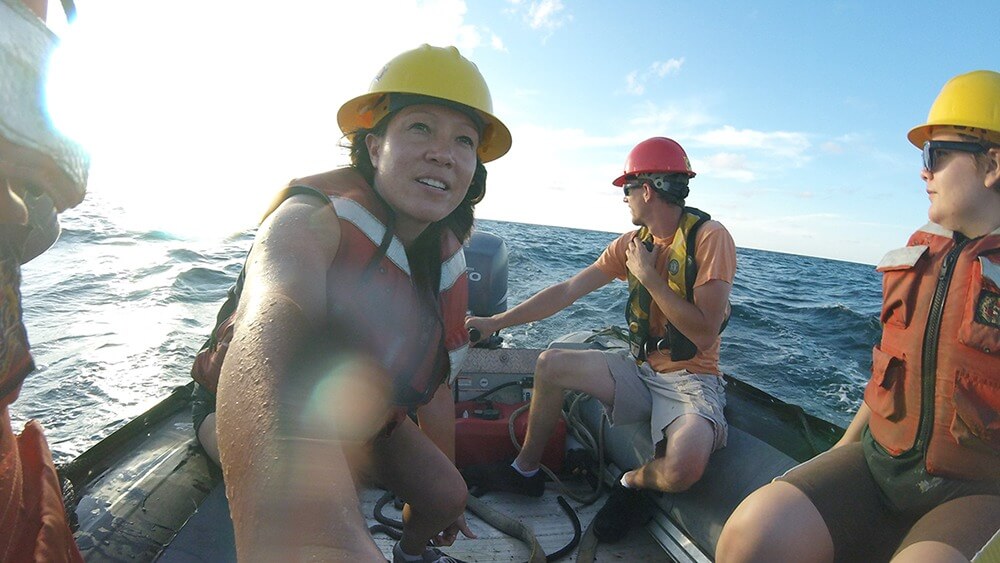 From left to right: Bright light from sun, ocean as background, 3 individuals on a boat: female with dark hair, yellow hard hat, orange vest, male with red hardhat, black sunglasses, orange tshirt, yellow vest and light shorts, person with yellow hard hat, orange vest, green shorts, and black sunglasses.