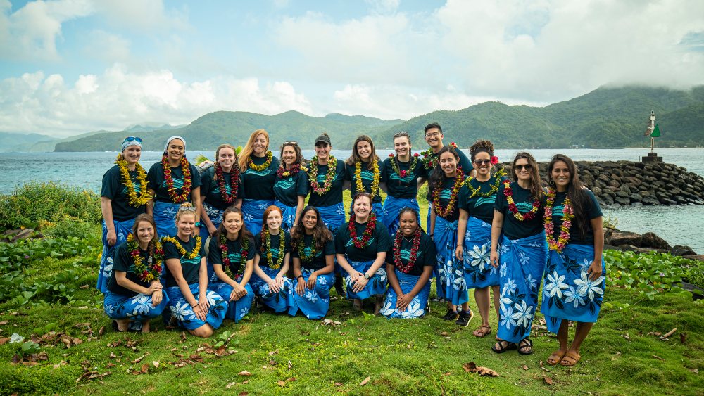 people wearing matching t-shirts, blue lavalavas, and fresh flower leis pose together for a photo in front of the ocean