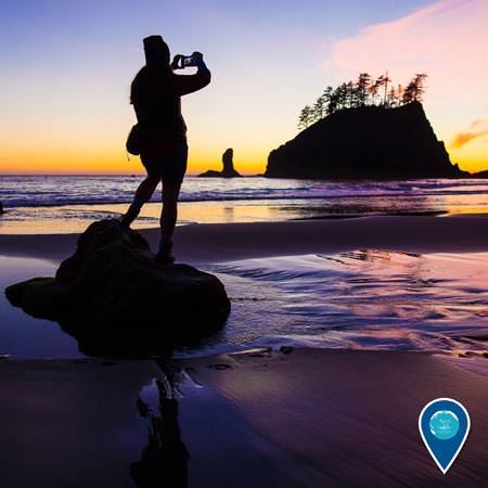 person standing on a rock on the beach taking a photo of the sunset with cannonball island in the back ground