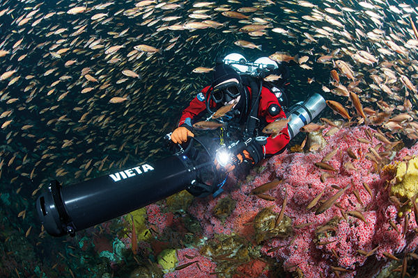 diver filming a reef while a school of fish swim around him