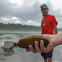 a person holds a razor clam up for the camera, while a man in the background stands by looking on the beach