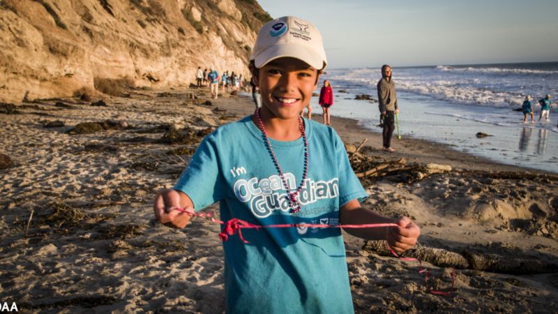 A young boy poses for the camera by holding a piece of trash up at a local beach cleanup. Other people cleaning up and the shore of the beach are seen in the background.