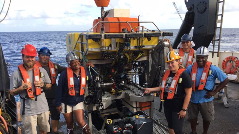 A NOAA crew consisting of 5 people all wearing hard hats and bright orange life vests surround an ROV (remotely operated vehicle). The blue sky meets the blue ocean at the horizon in the background.