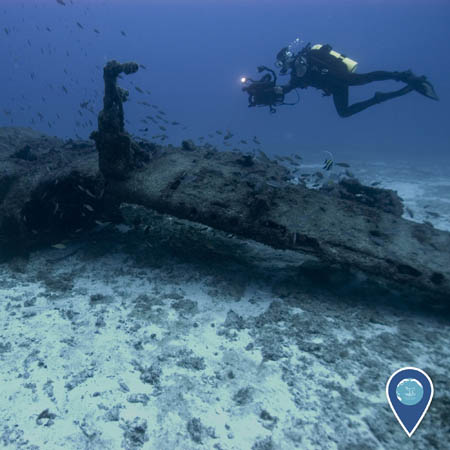 A diver carrying a camera swims above a sunken aircraft wing.