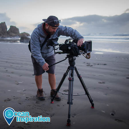 Nick stands on a beach, leaning over a video camera set up on a tripod. This image was taken on Ruby Beach in NOAA Olympic Coast National Marine Sanctuary, and the ocean and sea stacks are visible in the background.