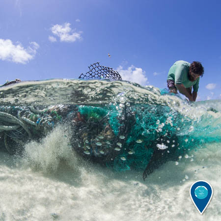 A person bends over a large conglomeration of fishing nets and other debris that is submerged in shallow ocean water.