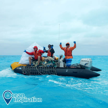 Three people on an inflatable boat holding their hands up in celebration; the boat is holding piles of nets and other debris.