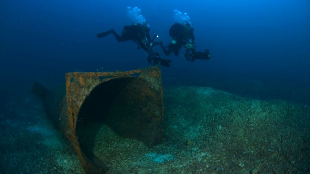 Two divers underwater observing pipes previously used for treasure salvage exploration and excavation during the Bright Bank Excavation.