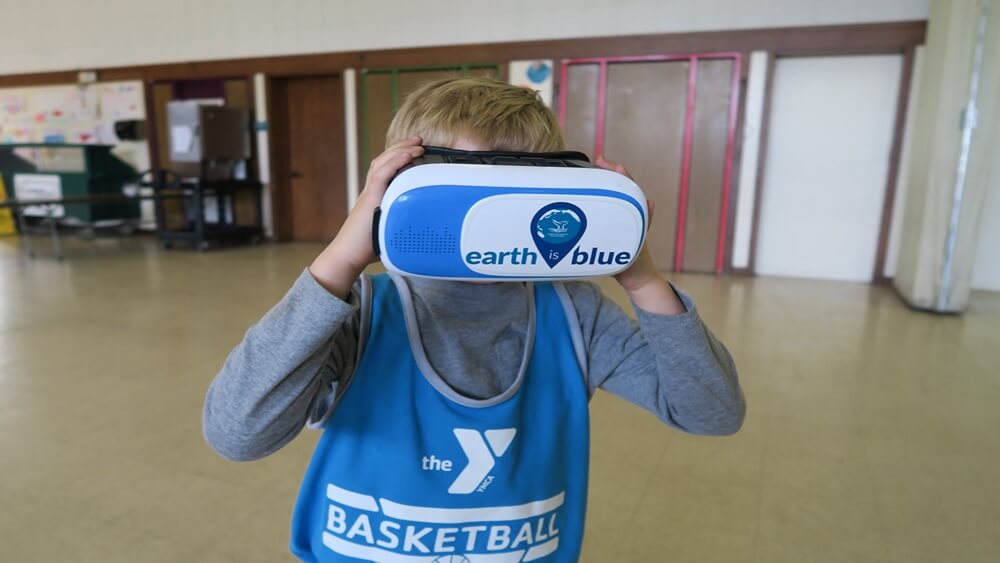 A young boy with blonde hair, a gray long sleeve shirt and a YMCA Basketball blue jersey, and a virtual reality headset on with the Earth Is Blue logo.