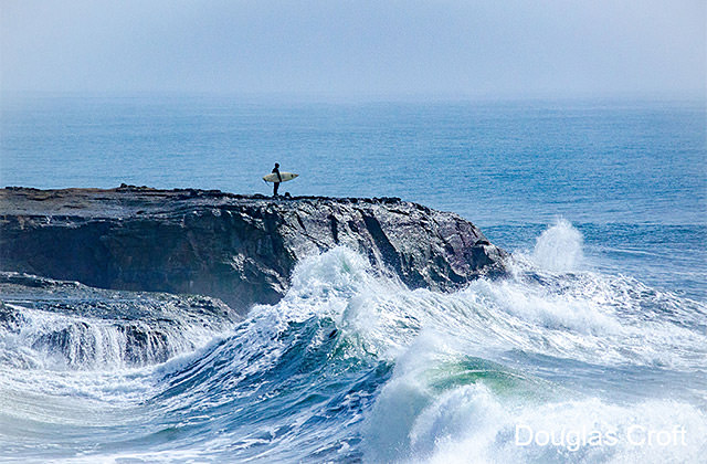surf standing on rock cliff while waves crash around the cliff