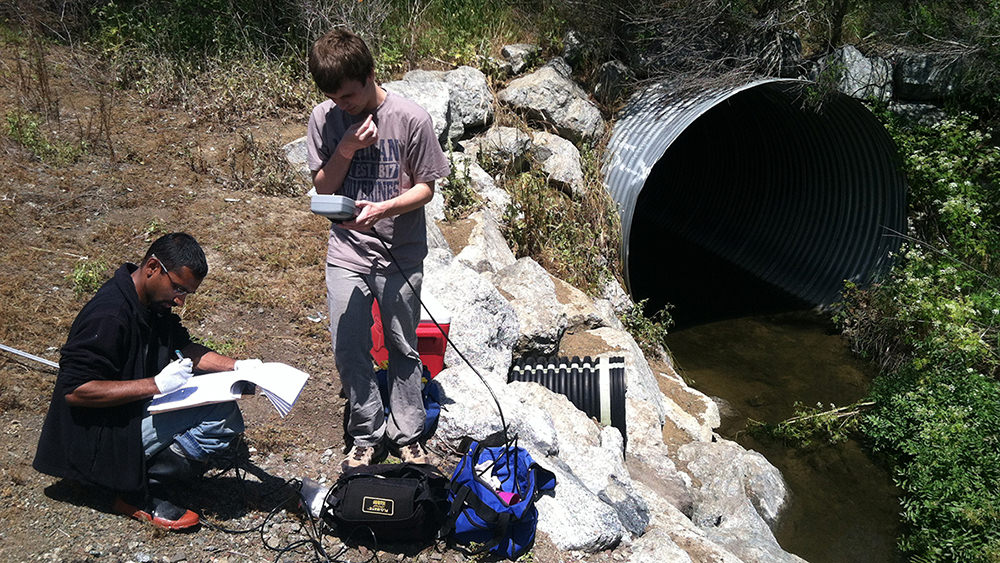 two people with monitoring equipment standing next to a culvert