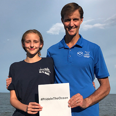 a man and a girl standing in front of the ocean holding a #prideintheocean sign