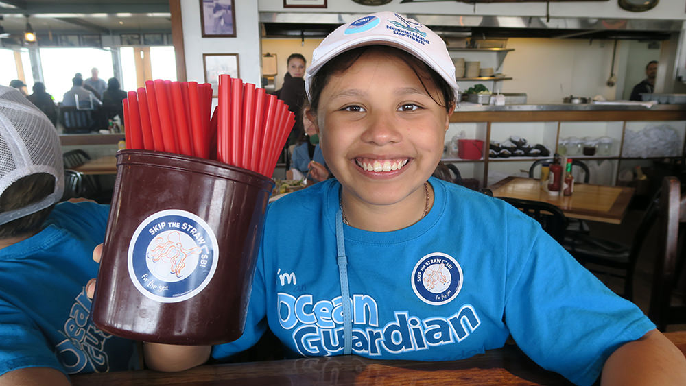 A young student holds up a container filled with red, plastic straws. She had placed a sticker on the side of the container that says “Skip the Straw”.