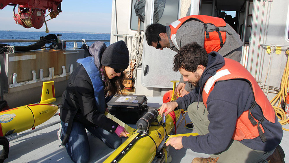 three people gathered around a yellow autonomous underwater vehicle on the deck of a ship