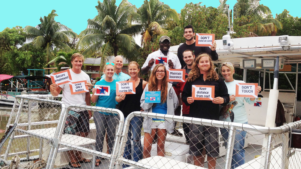 A group of people standing together on the back of a boat holding signs that read 'keep distance from reef' 'don't touch' 'leave only bubbles' 'secure equipment' 'dont stand on coral' 'don't harass marine life' 'maintain buoyancy'.