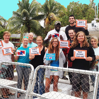 A group of people standing together on the back of a boat holding signs that read 'keep distance from reef' 'don't touch' 'leave only bubbles' 'secure equipment' 'dont stand on coral' 'don't harass marine life' 'maintain buoyancy'.