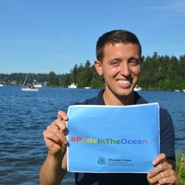 person smiling by water with a sign that reads #PrideInTheOcean