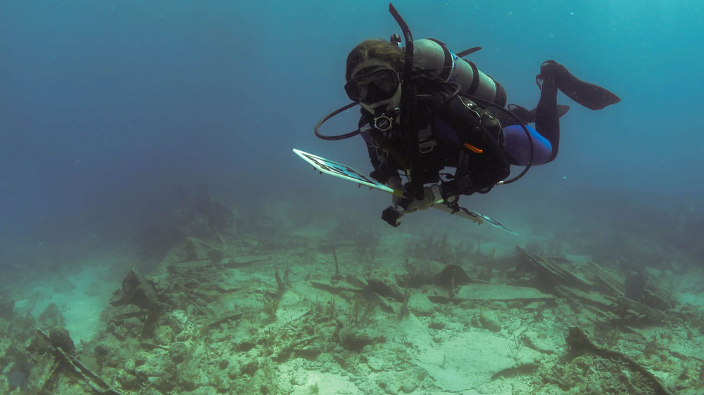 Diver swimming over a shipwreck while holding scientific equipment.