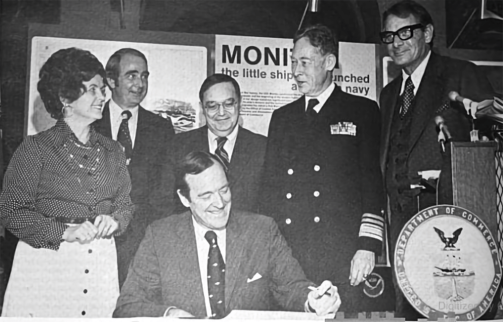 a black and white photo of people standing behind a man while he signs a document