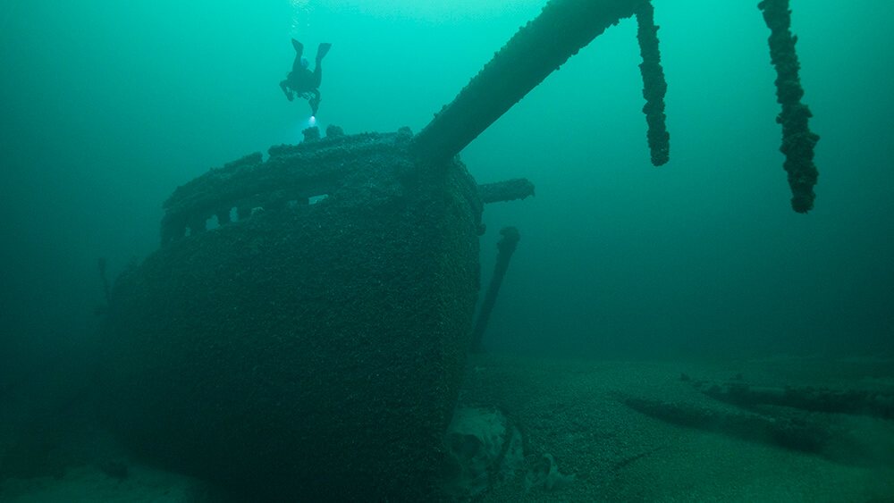 a shipwreck with a long bowsprit rests on the seafloor as a scuba diver epxlores it