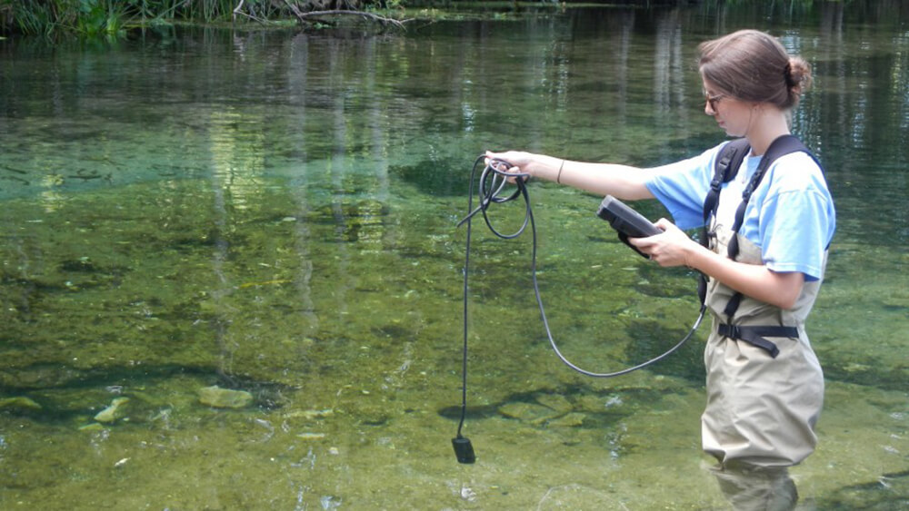 a researcher wearing waders stands in shallow water holding a sonde to take water quality measurements