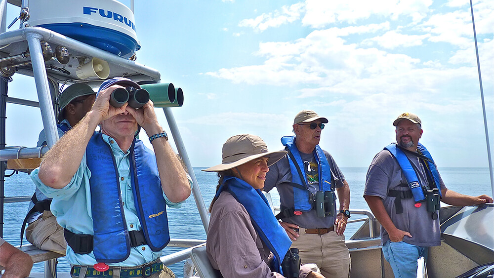 a volunteer peering through binoculars on a research vesselw with several other people aboard in the background