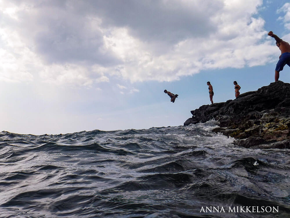 three people wait on the rocks, one dives into the water