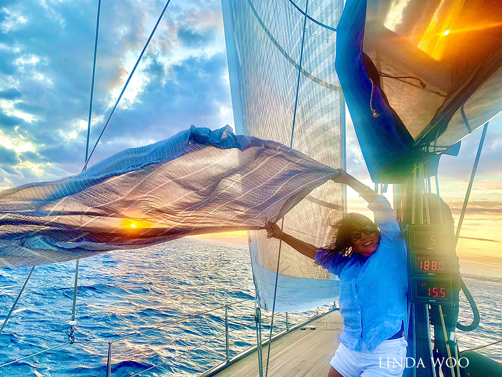 A person on a sailboat at sunset holding a shawl in the wind.