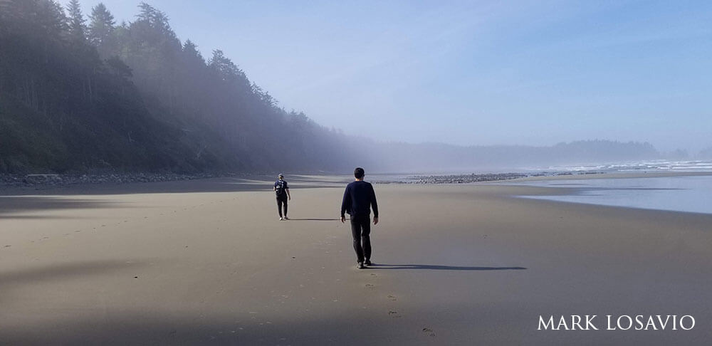 Two people on the beach, forest to the left, and misty ocean to the right