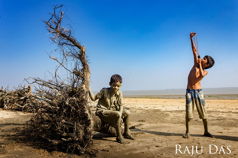 Two children sitting on the beach. One sits on a tree stump, the other holds a slingshot.