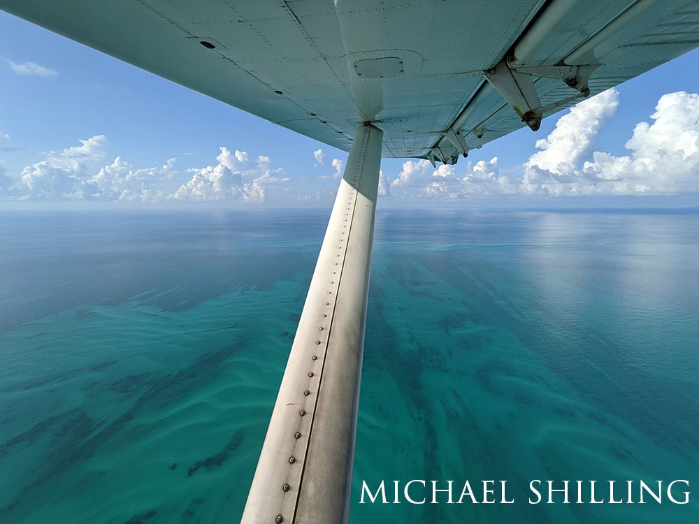 photo from an airplane, part of the plane visible. Bright, clear blue-green ocean below.