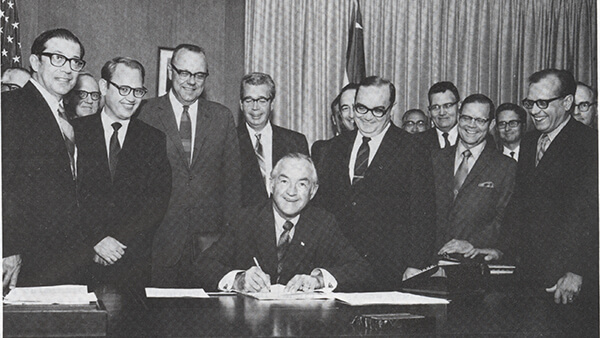 a black and white photo of people in business attire standing around a man signing a document
