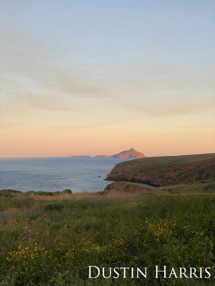 The sun sets on a cliff-side meadow overlooking islands in the distance.
