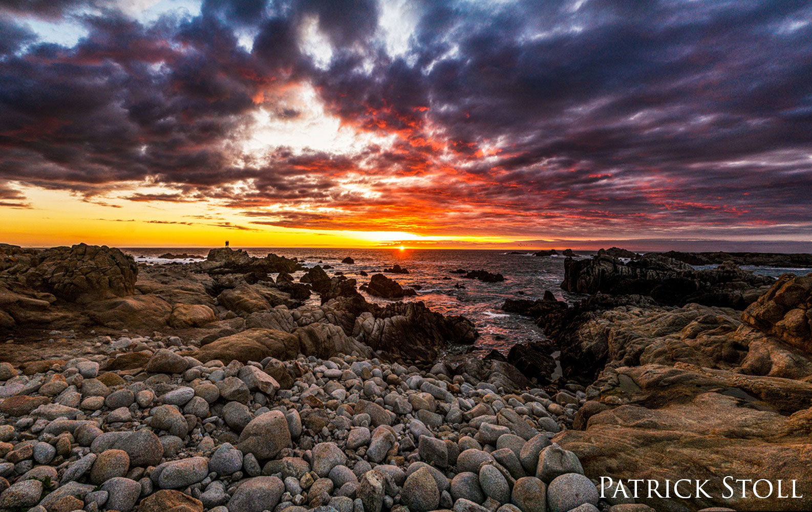 Two people watch the sunset on a rocky shore.