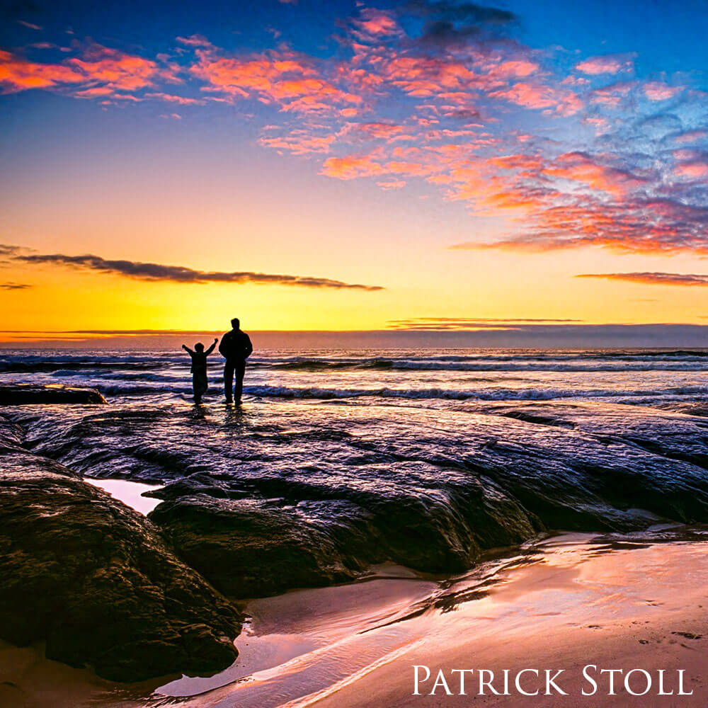 Man and boy watch the sunet atop a rock on a beach.