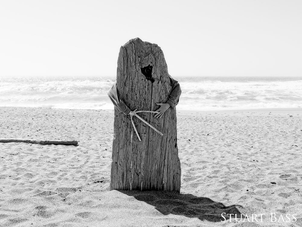 Man wearing a spooky mask wrapping arms around a plank in the sand, one of the man's hands is disguised as a branch. Photo is black and white. 