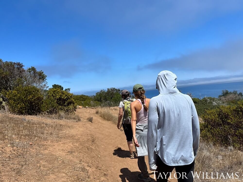 Three people hike on a trail with ocean in the distance.