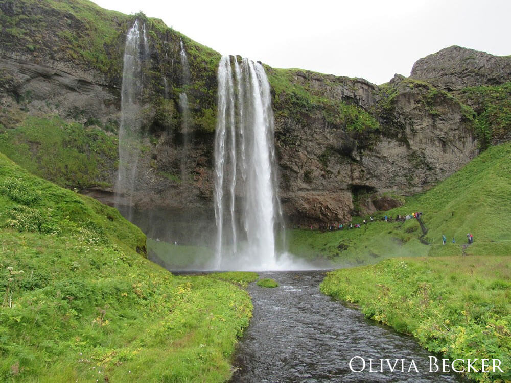 Giant waterfall with grass surrounding it, people are seen in the distance venturing under the waterfall.