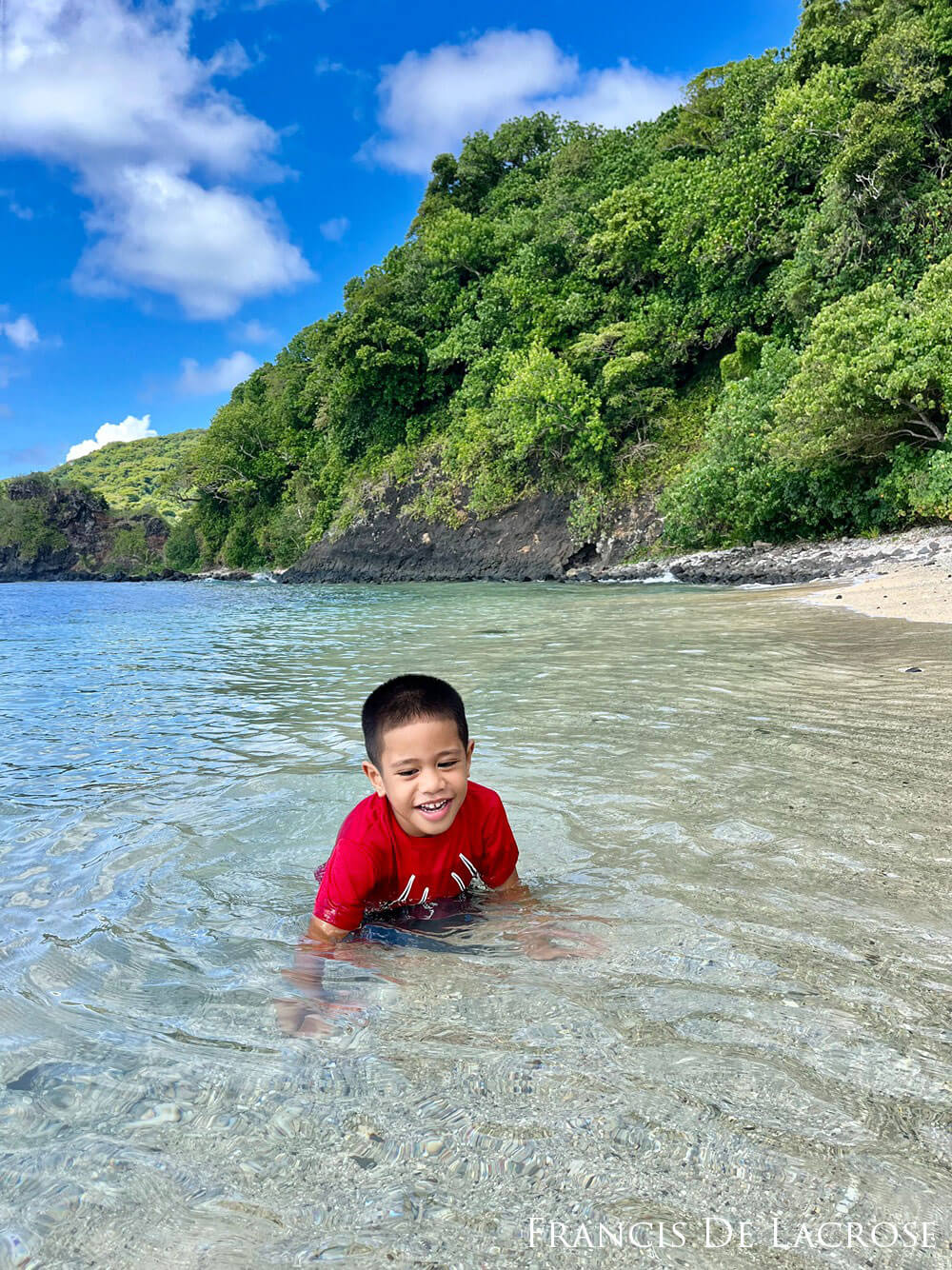 Child in the shallow, clear waters with green trees on a mountain in the background.