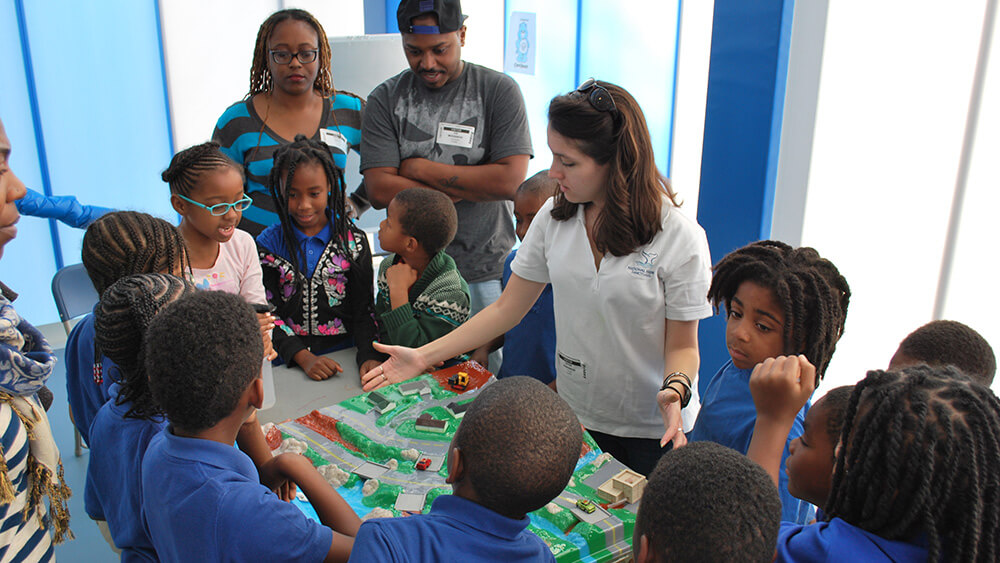 children gather around a docent who is using a large model of a watershed to teach about the impacts of pollution on aquatic ecosystems