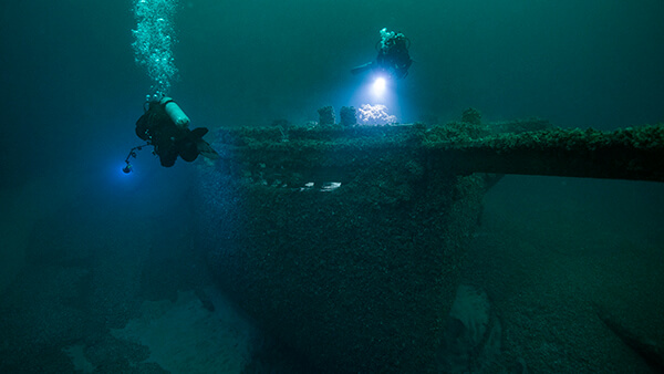 two scuba divers swim around a shipwreck in dark water. Their lights illuminate the shipwreck.