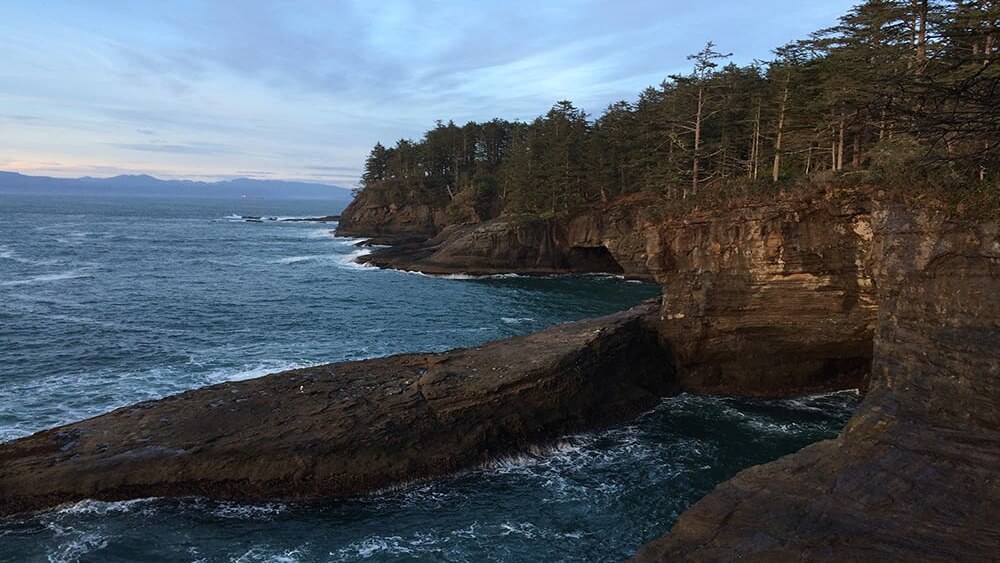 Hillside of Cape Flattery overlooking ocean and rocks below, tall trees on edge of the cliffs, and mountains on the horizon with a blue and pale orange sunset.