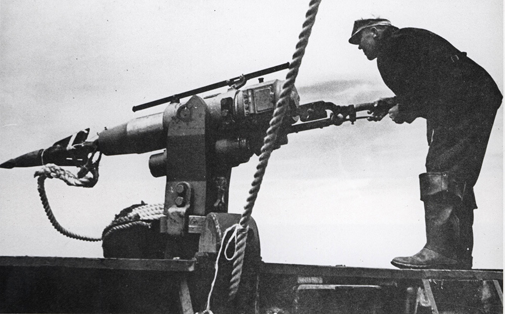 A photo from 1951 shows a man wearing a cap, jacket and pants tucked into rubber boots standing at a large whaling harpoon getting ready to fire it.