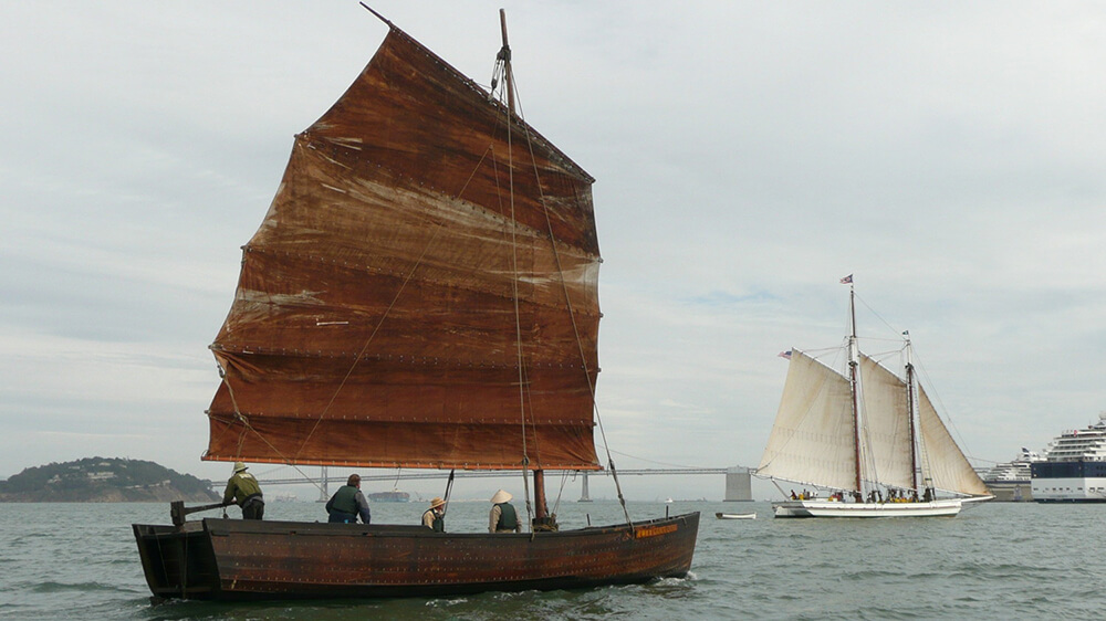 a dark wooden vessel on the water with a single sail fully raised