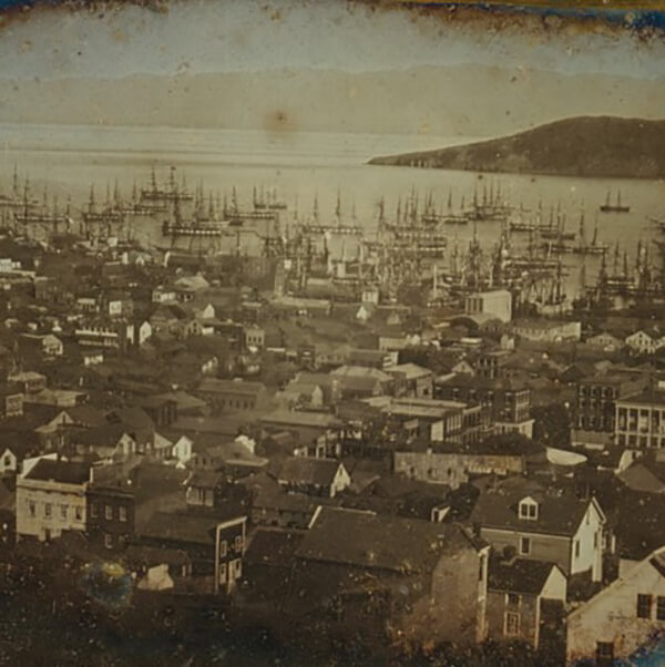a faded photograph showing a residential city with a bay in the background that has several sailing vessels on it