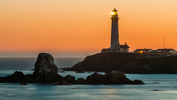 a lighthouse on a rocksy shoreline during a very orange sunset