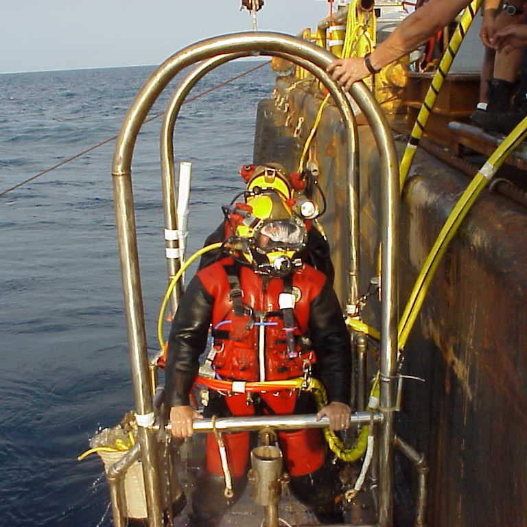 divers wearing metal helmets and full diving suits stand on a platform at the back of a ship