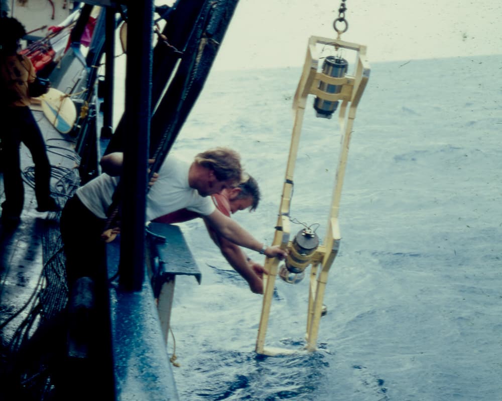 crew on a research vessel lean over the side of the vessel to lower a metal frame with cameras attached to it down into the water, assisted by a lift davit and cable system
