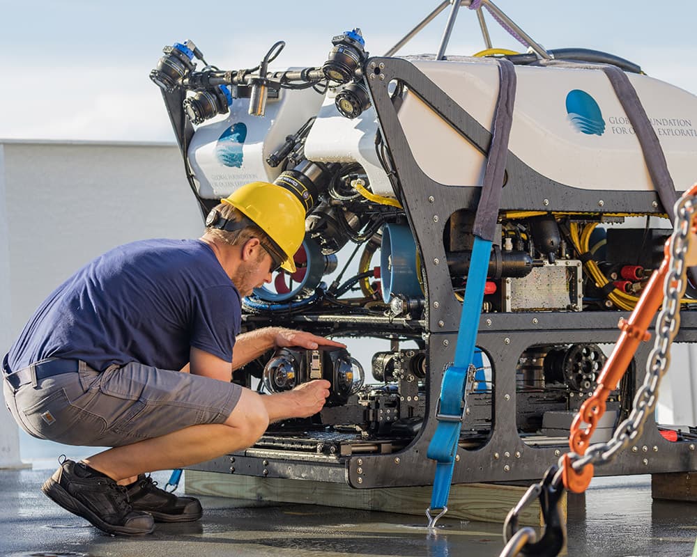 an engineer wearing a hard hat attaches cameras to a remotely operated vehicle sitting on the deck of a ship